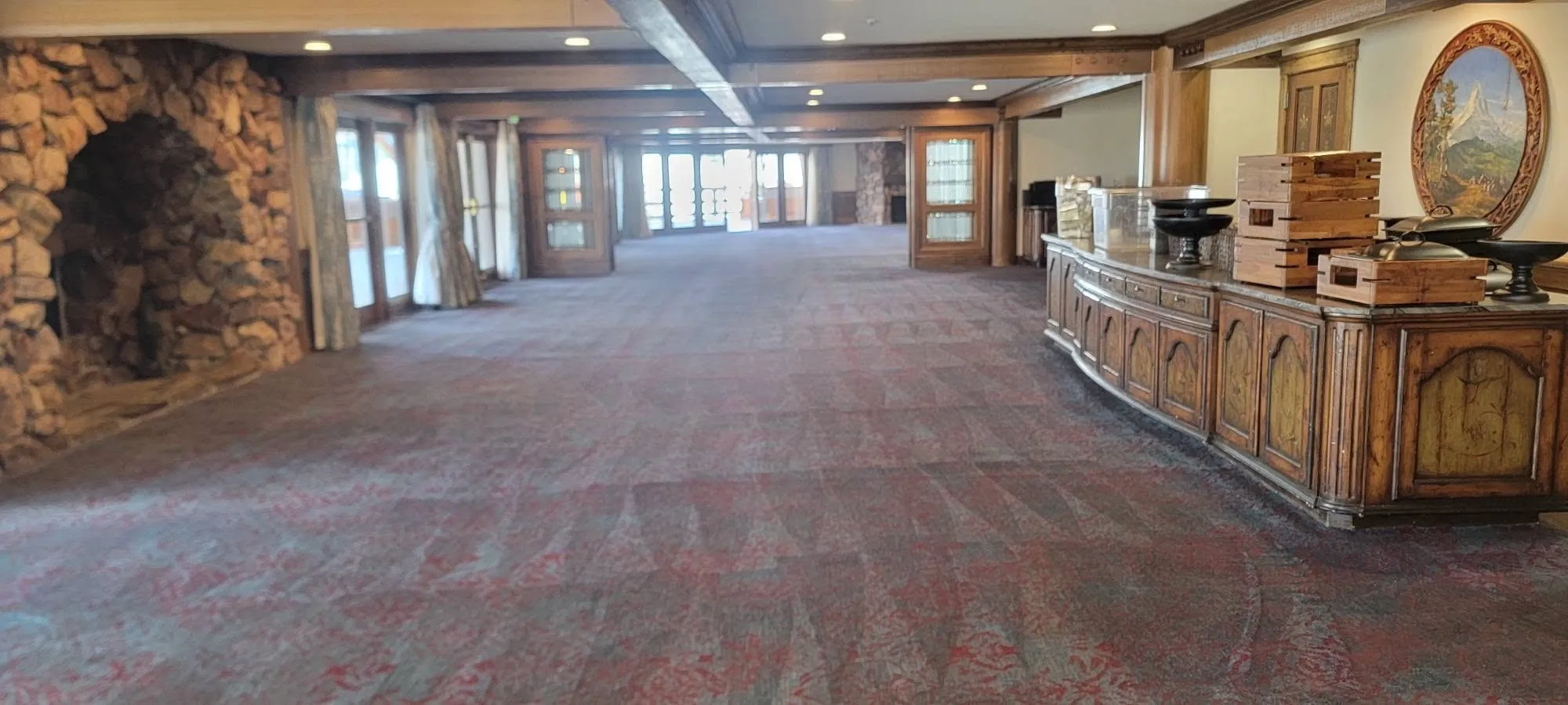 A wide lodge pre-function space with patterned broadloom carpet, a stone fireplace alcove at left and a carved wood buffet at right