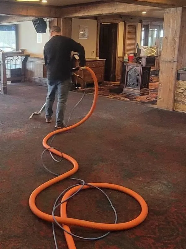 A Park City Carpets technician steam-cleaning patterned carpet in a timber-and-stone lodge, an orange solution hose looping across the floor behind him